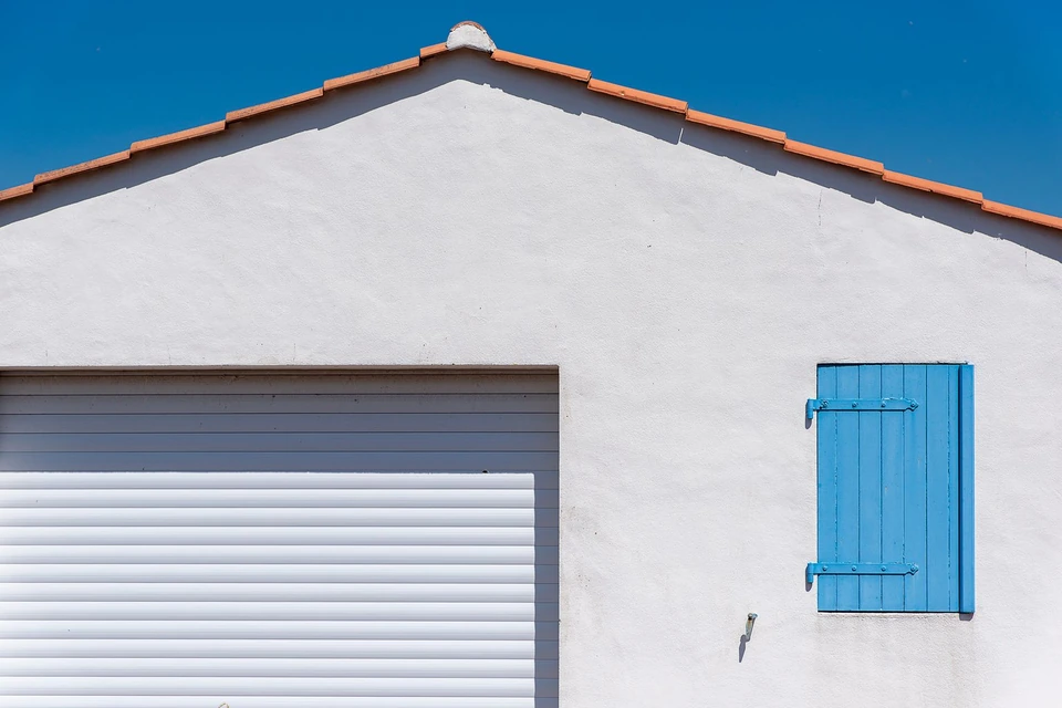 Pignon enduit blanc sous une rive de tuiles, avec porte de garage et volet bleu